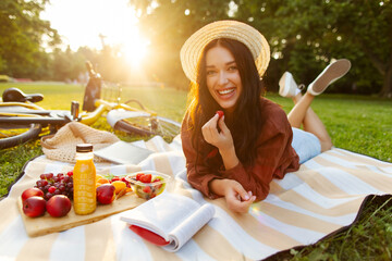 Beautiful young woman eating fruits and reading book while having picnic and lying on blanket in green park, chilling outdoors