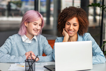 Smiling diverse young women collaborating on laptop in modern coworking space. Concept of teamwork, digital nomads, freelance, generation Z and creative business