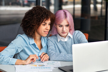 Young diverse women working together at laptop and analyzing business documents. Concept of teamwork, digital collaboration, mentorship and modern workplace