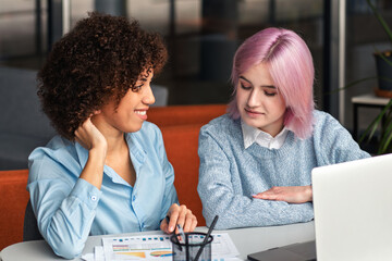 Young diverse women working together at laptop and analyzing business documents. Concept of teamwork, digital collaboration, mentorship and modern workplace