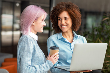 Two smiling young women having friendly conversation while holding laptop and coffee in modern workspace. Concept of networking, teamwork and female empowerment