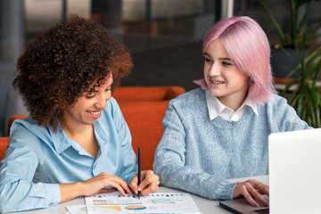 Smiling diverse young women collaborating on laptop in modern coworking space. Concept of teamwork, digital nomads, freelance, generation Z and creative business