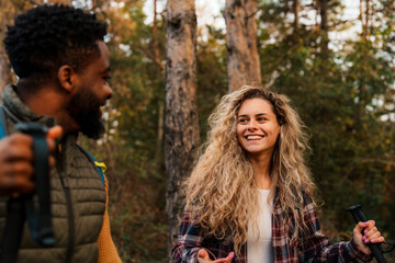 Two diverse people hiking in a forest, they are in conversation, carrying trekking poles and wearing backpacks.