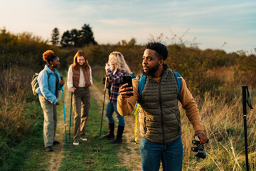 A black man hiker holds binoculars and checks map on phone as dusk sets in. Three friends chat nearby in the green surroundings, enjoying their adventure in nature.