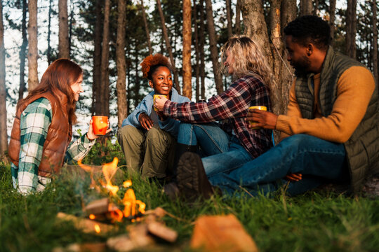 Diverse friends enjoying camping evening around campfire, drinking hot beverages and sharing genuine moments in pine forest. Authentic outdoor lifestyle. - Powered by Adobe