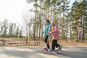 Little girl riding scooter while walking with her grandmother in the forest on a sunny day