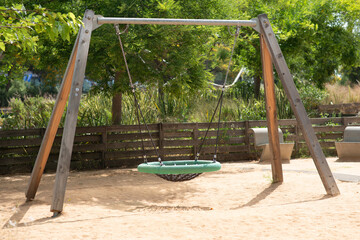 Modern playground featuring a nest swing with a round woven seat, suspended by ropes from a wooden and metal A-frame. The swing stands on a sandy surface. Ideal outdoor recreational space for children