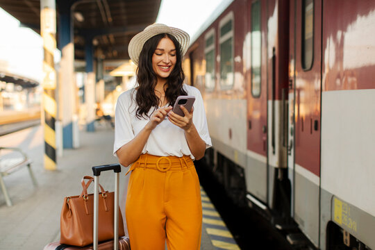 Positive young lady traveler using her smartphone while standing at train platform. People and transportation concept