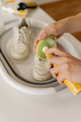 Vertical shot of man cleaning pair of dirty white shoes in foldable basin using brush and cleaning foam, taking care of footwear hygiene and maintenance, close-up. Leather footwear care concept.