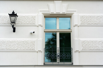 Modern white building facade with classic decorative elements, vintage street lamp, security CCTV camera and window with reflection and metal balcony railing on a sunny day. Urban architecture detail.