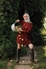 A fashionable woman in a red dress is holding a bucket full of apples. The model is posing in a...