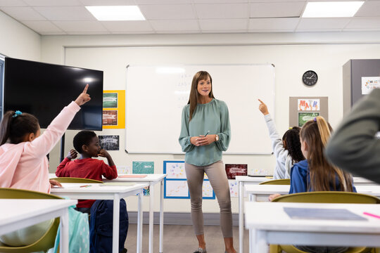 Female teacher engaging with diverse students in classroom, children raising hands, at school