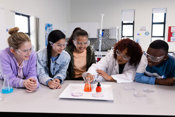 In school, diverse students wearing goggles observing colorful chemical reaction with female teacher