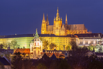 Illuminated Saint Vitus Cathedral and surrounding buildings in Prague Czech Republic at night with gothic towers and Baroque church in foreground