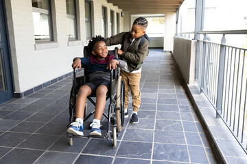Happy boy pushing friend in wheelchair along school corridor, enjoying friendship