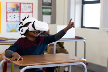 African american boy using vr headset in classroom, enthusiastically raising hand, at school