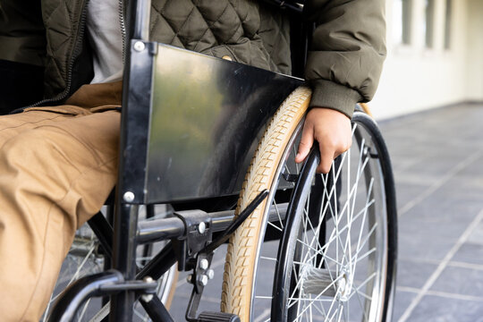 African american boy in wheelchair moving forward, wearing warm jacket and brown pants, at school