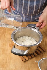 Cooking boiled rice on light wooden background. Woman pouring water from measuring jug over washed rice in metal pan. Cook's work, cooking.
