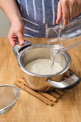 Cooking boiled rice on light wooden background. Woman pouring water from measuring jug over washed rice in metal pan. Cook's work, cooking.