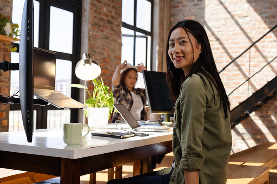 Asian woman coding on laptop in modern office, smiling confidently at camera