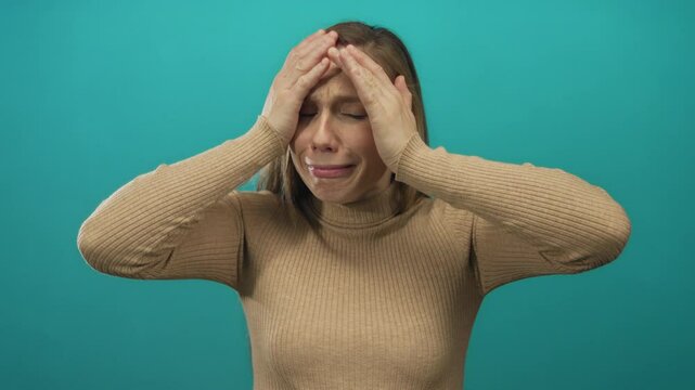 Young blonde woman showing frustration against an isolated green background wall with hands on head and distressed expression vividly illustrating stress and tension.