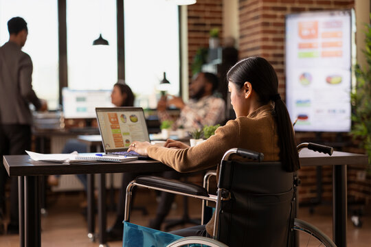 Professional woman in wheelchair using her laptop in brick wall office, catching up on work after a car accident. Asian female entrepreneur reviewing business project presentation in modern workspace. - Powered by Adobe