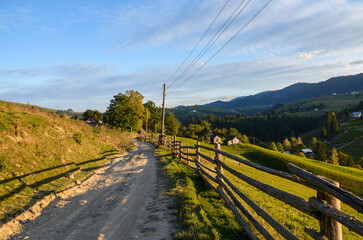 Peaceful dirt road stretches through a rural landscape, bordered by wooden fences and rolling...