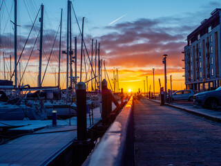 A sun rise over a marina with silhouettes of masts of many boats and yachts docked. Teal and orange...