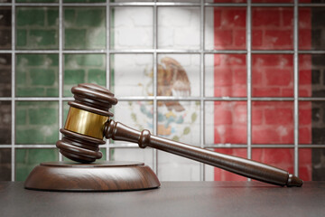 Wooden Gavel Resting on Table In Front of Prison Bars and A Brick Wall with Mexican Flag.