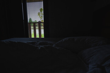 Dark moody bedroom interior with unmade bed and open blinds revealing a scenic terrace and greenery...