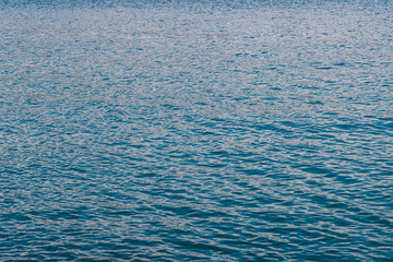 A small boat floats on the calm blue waters near the coast of Gaeta, with mountains and clouds in the background on a bright day
