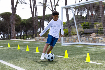 Multiracial boy child dribbling soccer ball through yellow cones on turf field with goalpost
