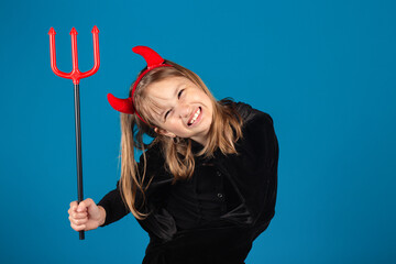 Funny child girl fooling around in devil halloween costume or mantle with horns and trident on blue...