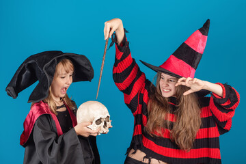 Halloween Theme. Young witch girl in striped sweater and black hat holding magic stick and making spell to the skull in another young sorceress hands against blue background