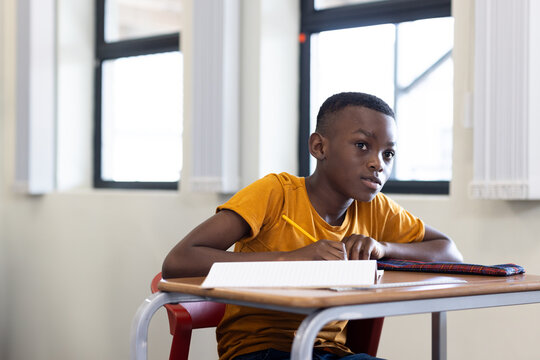 Writing African American boy holding yellow pencil over notebook at desk in classroom, copy space