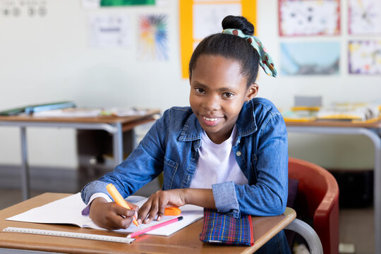 Sitting girl student writing in open notebook at wooden classroom desk, with orange highlighter