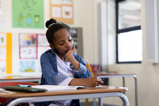 Writing girl leaning head at classroom desk, with pencil, open notebook, pencil case