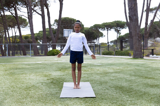 Child girl standing barefoot on gray yoga mat on fenced outdoor sports turf, with basketball hoop