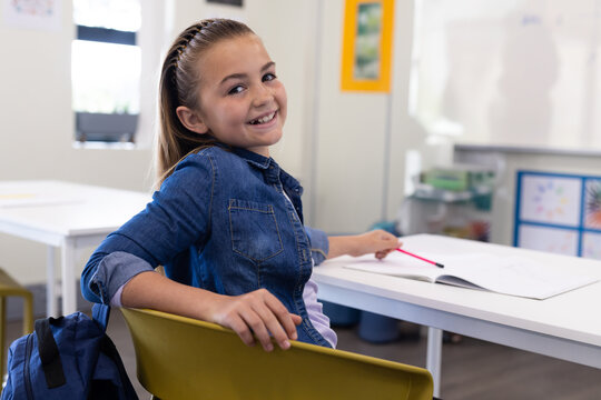 Female child student sitting at classroom desk turning toward camera while holding pink pencil