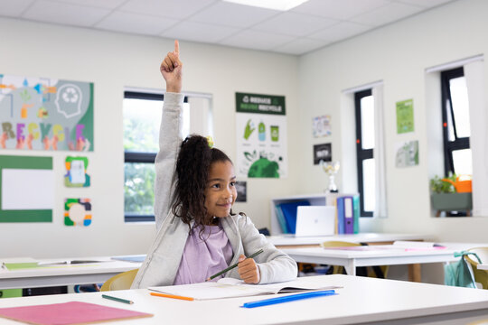 African American girl raising hand at classroom desk with recycling posters and holding notebook