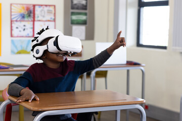 Student wearing white VR headset pointing at empty space at desk in classroom with laptop, posters