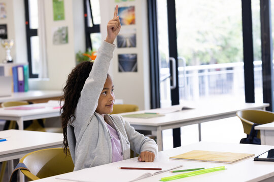 Female child raising hand and sitting at school desk with notebook pencils posters near glass door