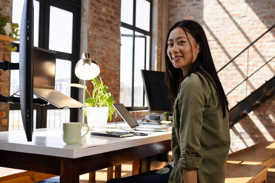 Chinese woman working at loft-style office in olive green shirt with dual monitors mug, copy space