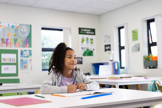 African American girl student sitting at classroom desk holding pencil poised over open notebook