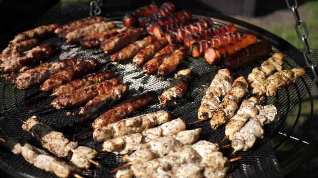 Close-up of grilled chicken, sausages, and meat skewers sizzling over an open outdoor flame. Smoke rises as fire flickers, all in warm natural sunlight.