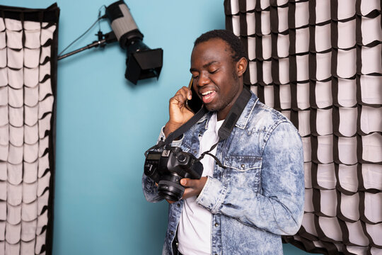 Black photographer on phone call, holding his DSLR and standing near his studio equipment. African American man with mobile device to his ear, wearing a camera around his neck and reviewing pictures.