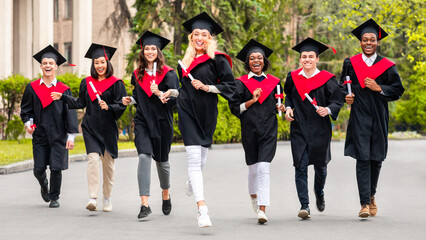 Delighted multiracial students guys and young ladies in graduation costumes running towards camera by university campus, holding diplomas and smiling. College, university graduation, education concept