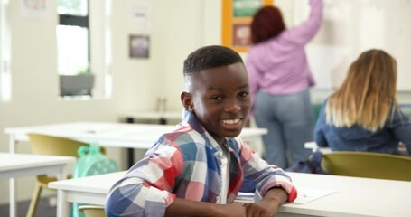 Smiling african american boy in classroom looking at camera while female teacher writes, at school