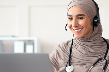 Cheerful muslim lady doctor having video chat with patient, using headset and laptop, talking to...
