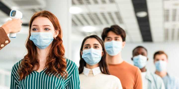 Morning At Work During Covid-19 Epidemic. Person with contactless laser gun thermometer measures body temperature of multiracial employees in protective face masks at entrance of modern office